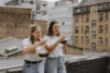 Zwei junge Frauen essen auf einer Terrasse an ein Geländer gelehnt im stehen
